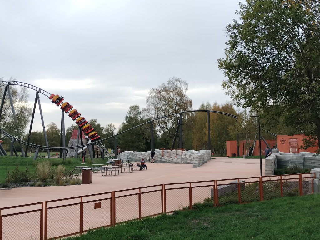 montagne russe Namazu Vulcania en fond
place avec tables et bancs devant
herbe bordée par une barrière au premier plan